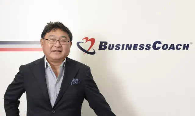 会社のロゴをバックに写真に写るビジネスコーチ代表取締役社長の細川馨氏