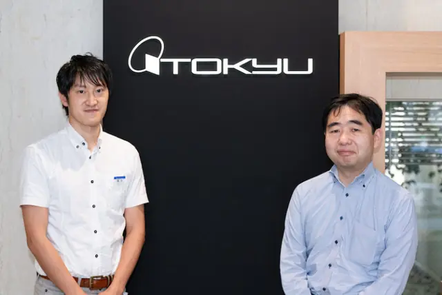 株式会社東急百貨店男性社員２人が会社のロゴの前に立っている