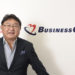 会社のロゴをバックに写真に写るビジネスコーチ代表取締役社長の細川馨氏
