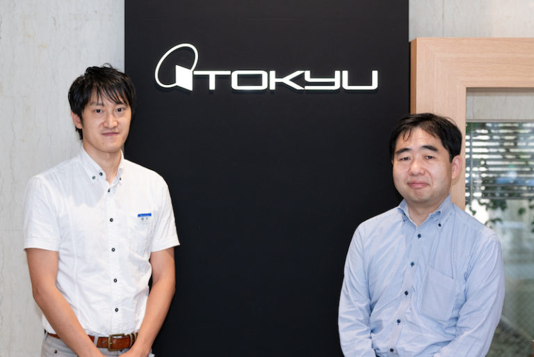 株式会社東急百貨店男性社員2人が会社のロゴの前に立っている